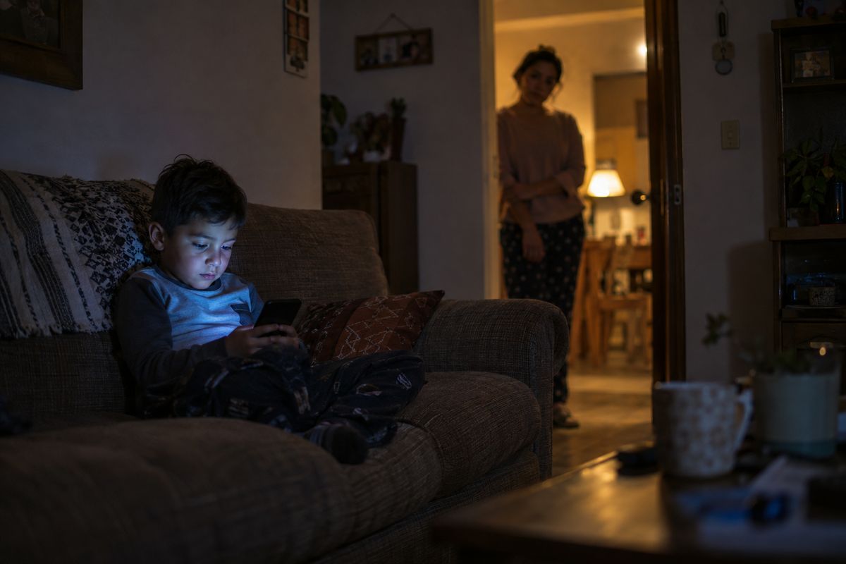 Un niño solo en el sofá con el celular; la mamá lo mira desde la puerta